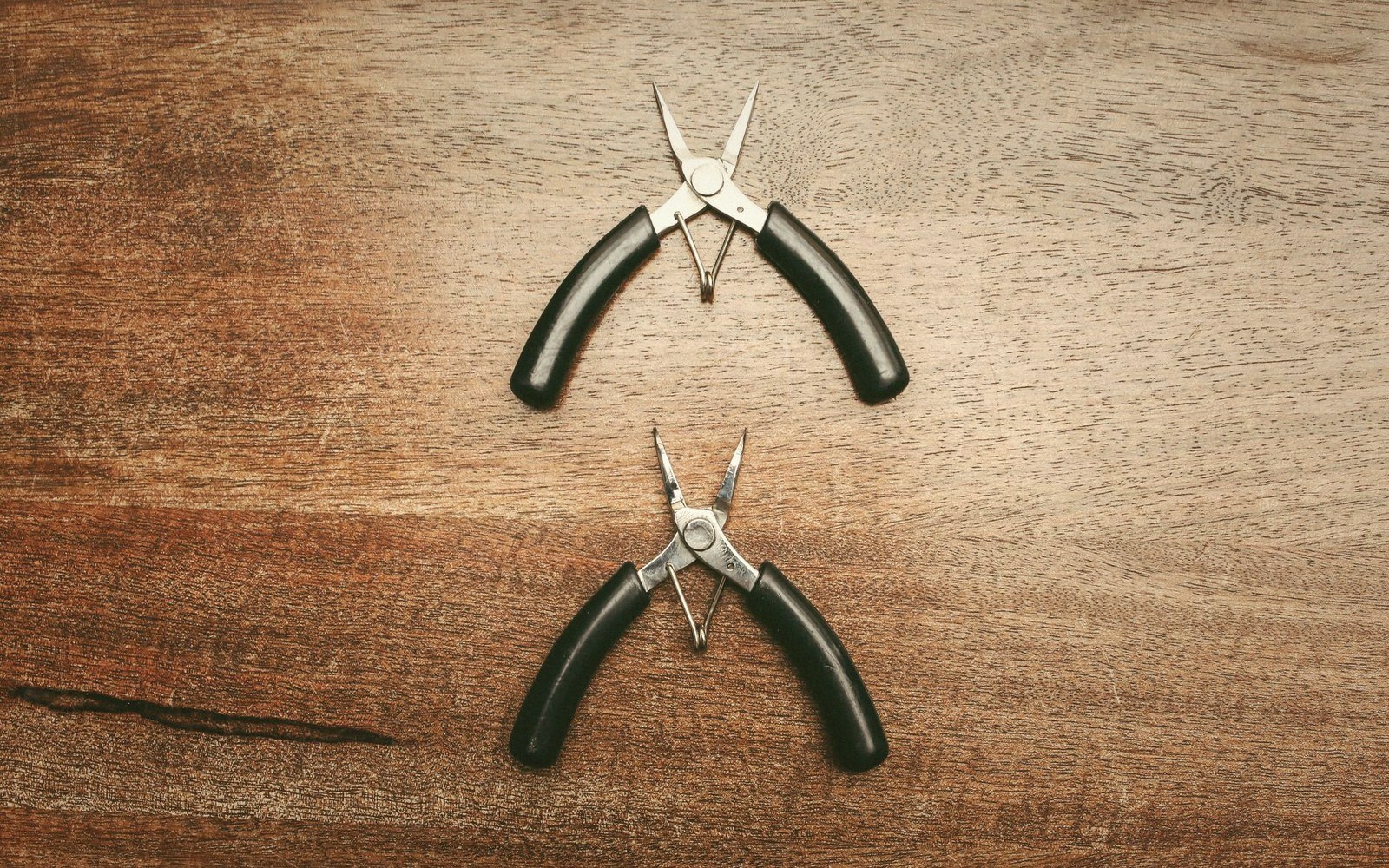 Two pairs of small jewelry pliers with black handles lie on a wooden surface